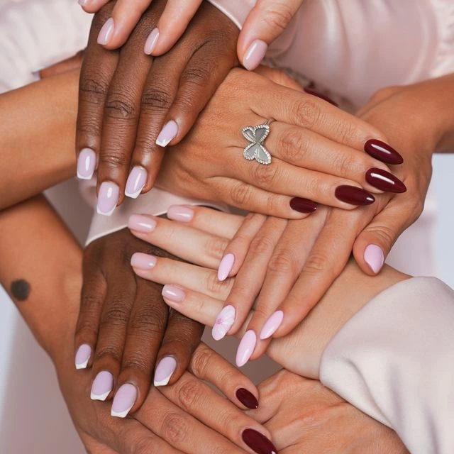 A group of hands with different skin tones are stacked together, each showcasing neatly manicured nails in shades of pink, nude, and deep red. One hand features a silver butterfly-shaped ring. The image highlights diverse nail designs and colors.