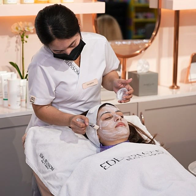 A spa therapist wearing a mask applies a white facial mask to a woman lying on a treatment bed. The woman is covered with a towel and has a headband holding her hair back, while the therapist uses a brush and a small jar of product. The setting appears to be a clean, modern beauty spa with skincare products on the counter behind them.