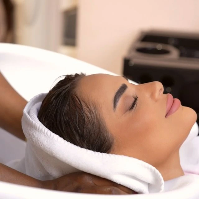 A woman reclines with her eyes closed at a salon hair-washing station. Her wet hair is wrapped in a white towel while a stylist supports her head. She appears relaxed, with soft makeup and a peaceful expression.